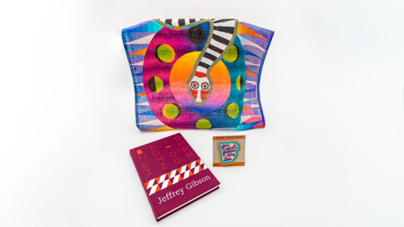 Bright colored tote bag with a snake on it next to a book and a patch on a white background.