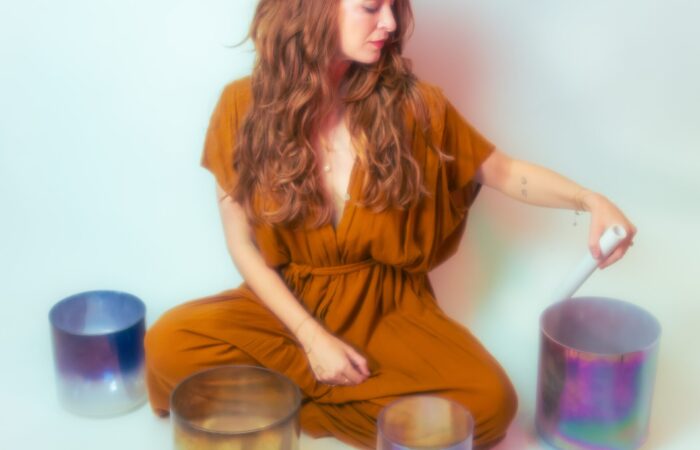 A woman in a rust colored jumpsuit sits on the ground, surrounded by different colored glass jars.