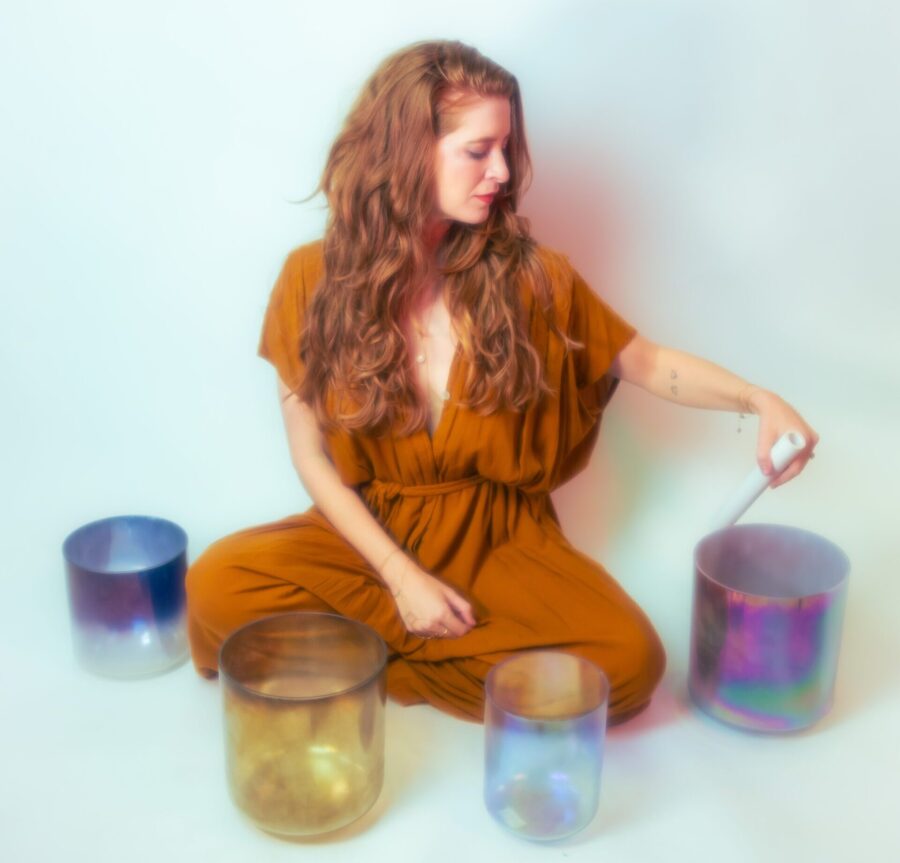 A woman in a rust colored jumpsuit sits on the ground, surrounded by different colored glass jars.