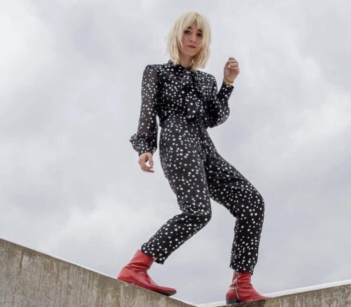 A woman in a black polka-dot jumpsuit with red boots stands on a concrete ledge.
