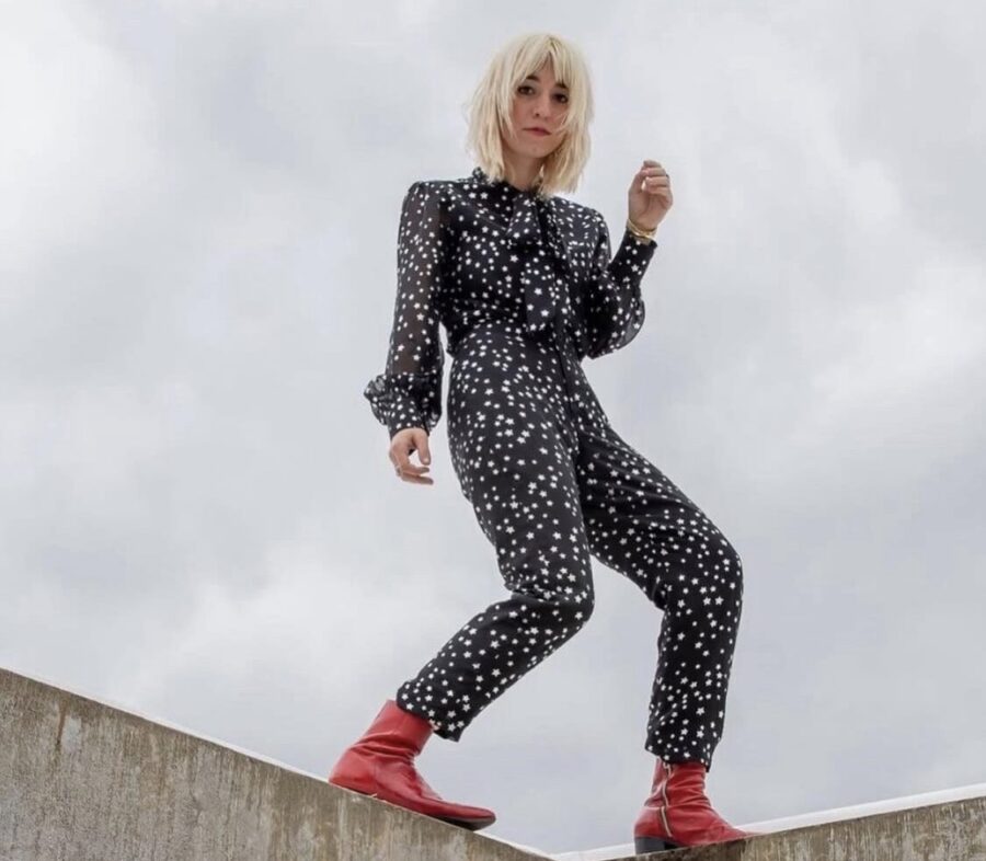 A woman in a black polka-dot jumpsuit with red boots stands on a concrete ledge.