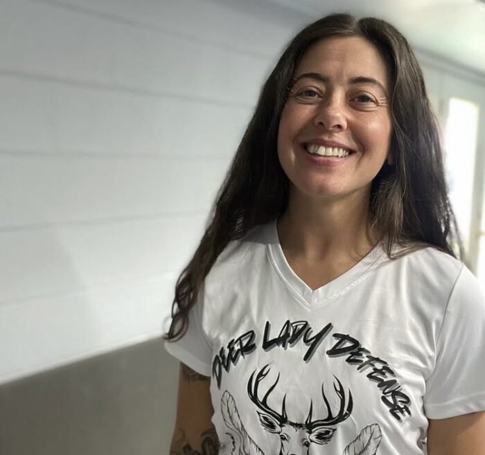 A woman smiles wearing a white shirt that says Deer Lady Defense.