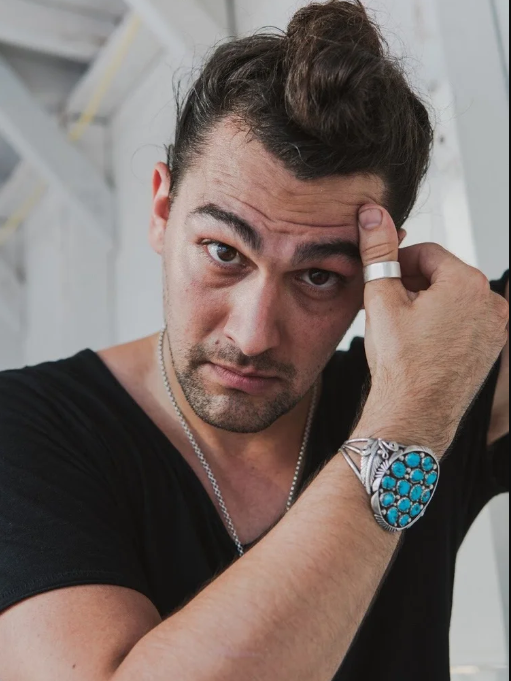 A man with a bun and silver jewelry poses with his hand on his forehead. 