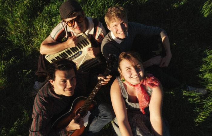 A group of four young people look up at the camera from a field with instruments.