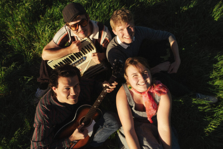 A group of four young people look up at the camera from a field with instruments.