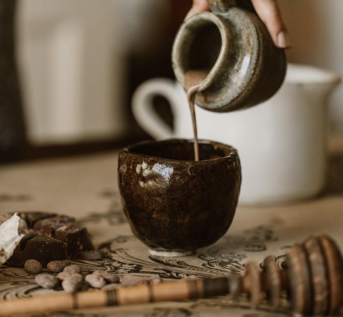 Drinking chocolate is being poured from pottery vase into another ceramic mug.