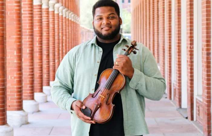 A man holds a violin in an outdoor hallway with brick columns.