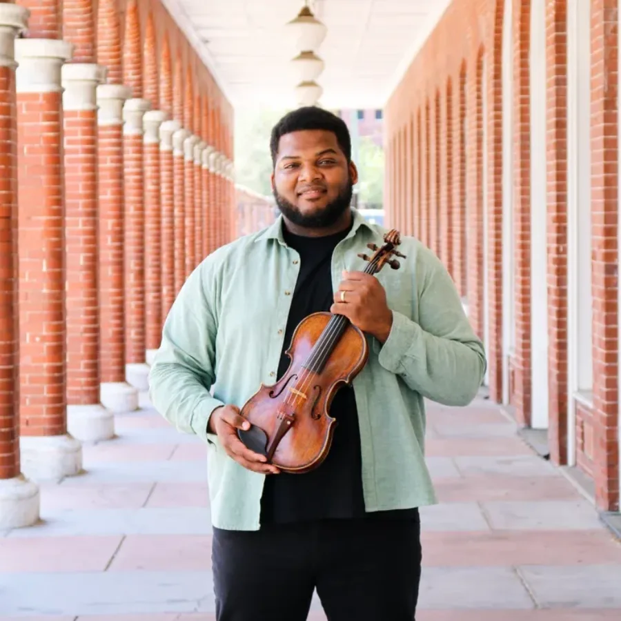 A man holds a violin in an outdoor hallway with brick columns.
