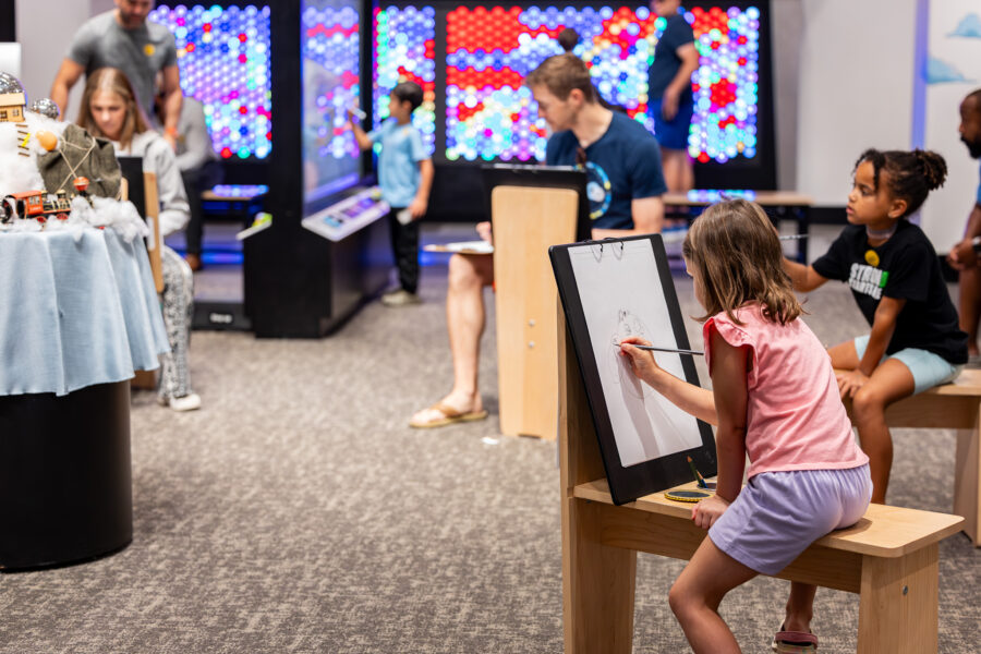 A group of adults and kids draw around a still life set up in an interactive art exhibit.