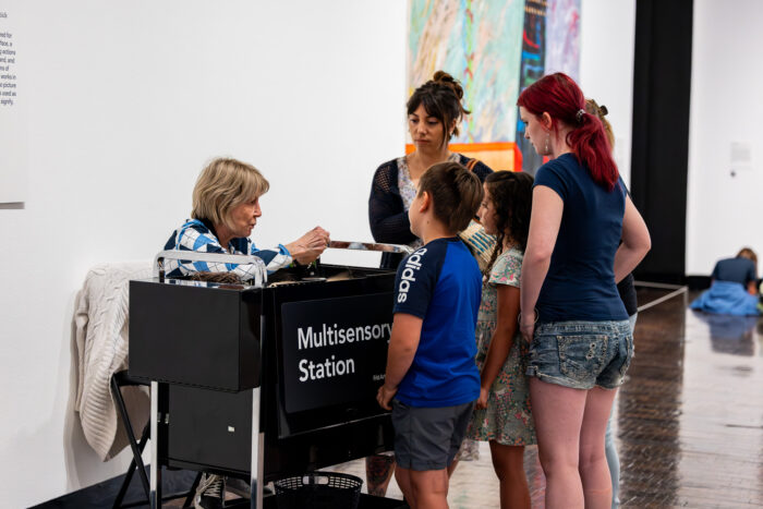 A group of adults and kids stand around a Multisensory Station in an art gallery.