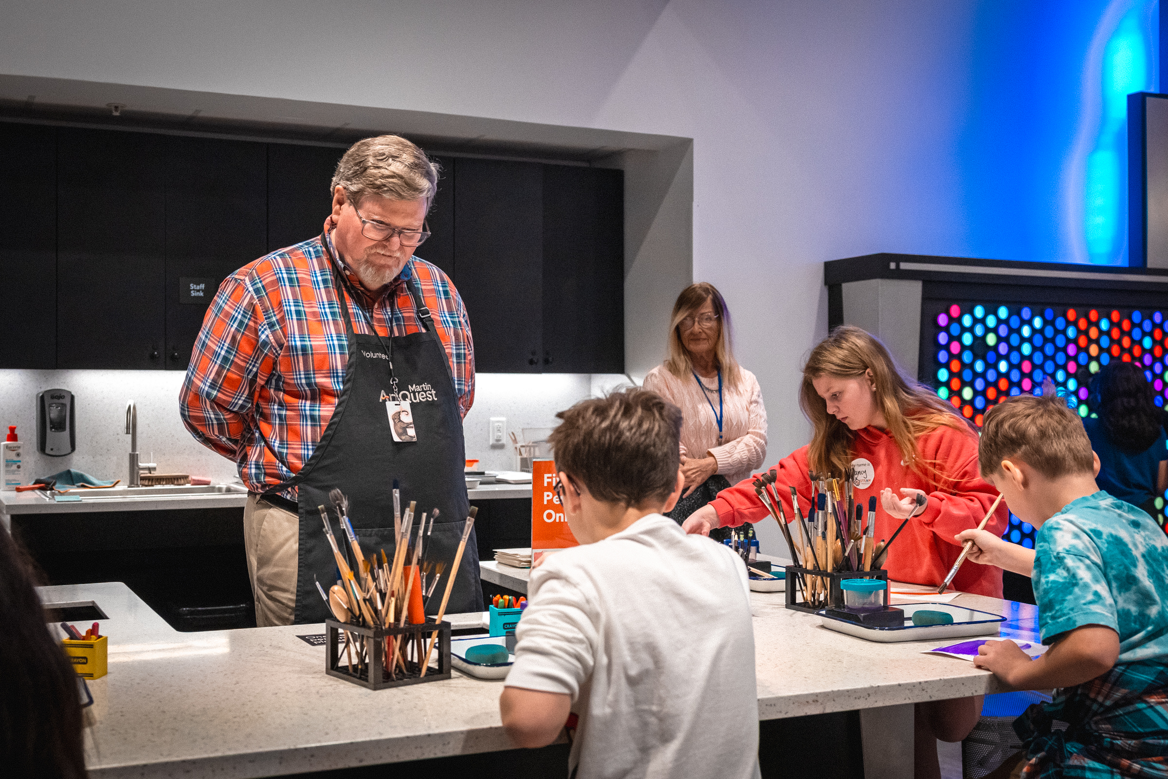 Man watching children make art and wearing a black volunteer apron
