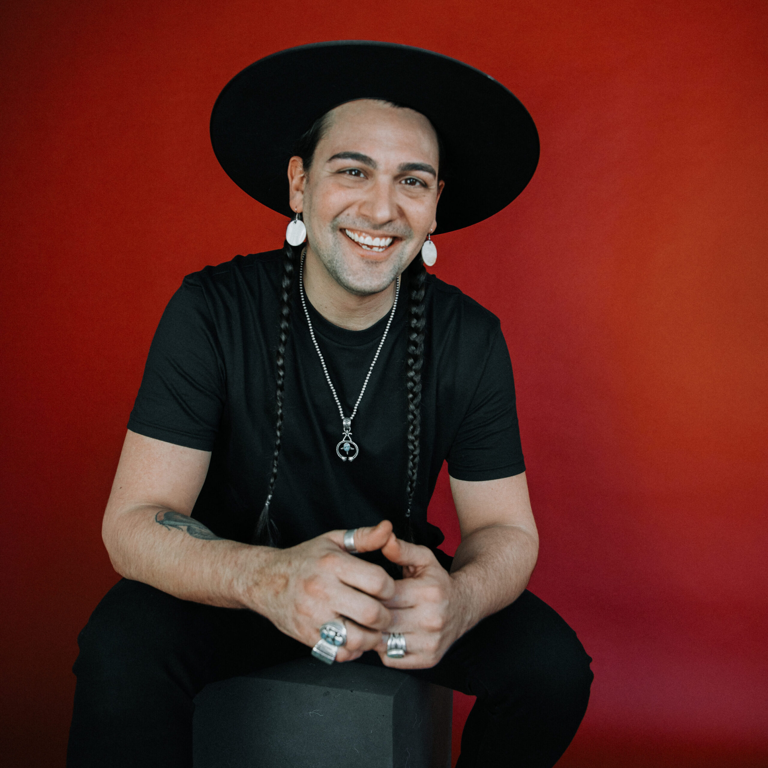 A man with two long braids poses in front of a red background with a black hat. 