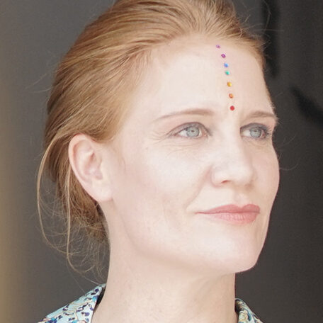 A woman with red hair poses with rainbow jewel dots on her forehead.
