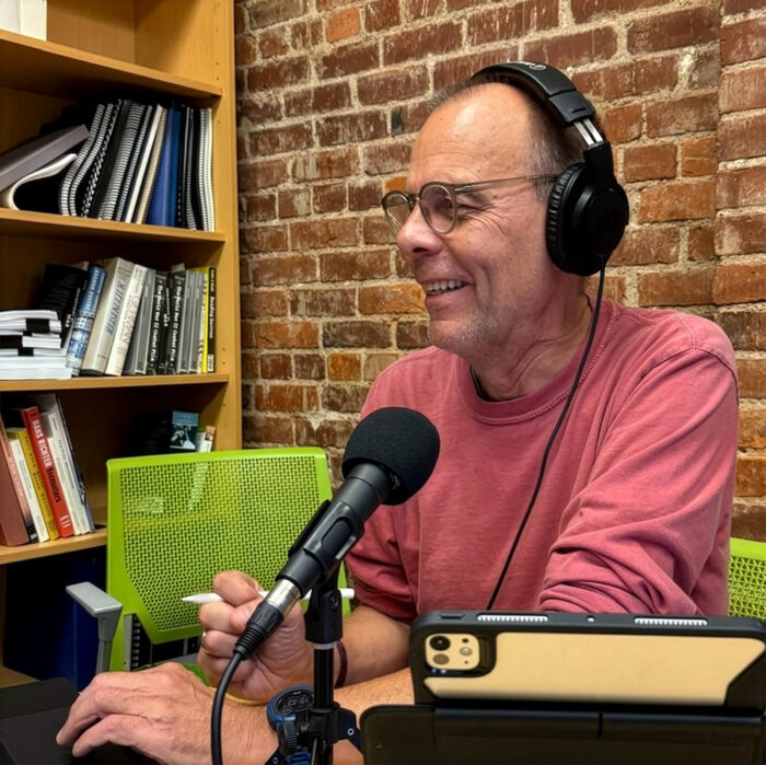 A man sits at a microphone with headphones in a room with brick walls.