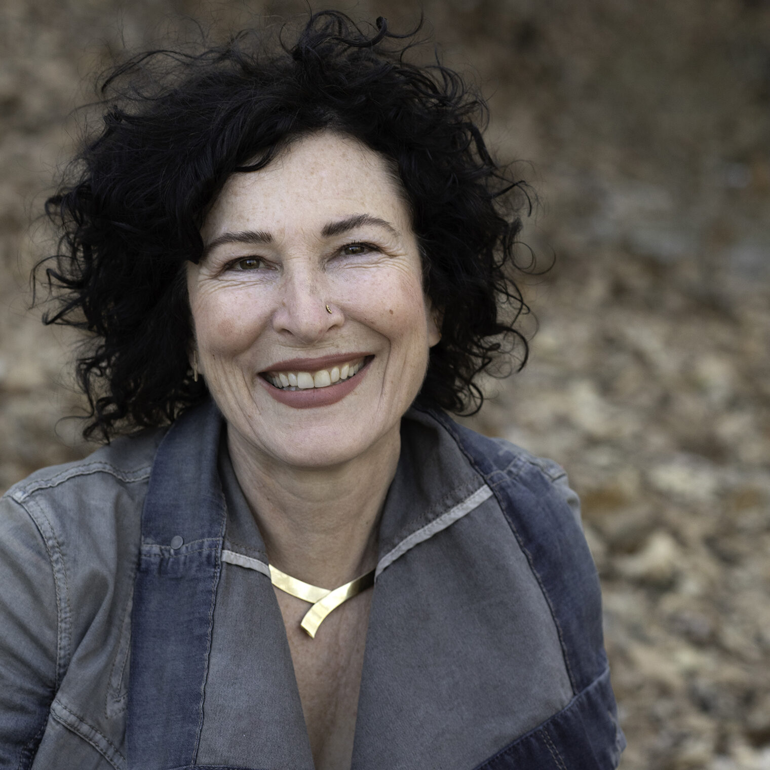 A woman with black curly hair smiles with leaves in the background.