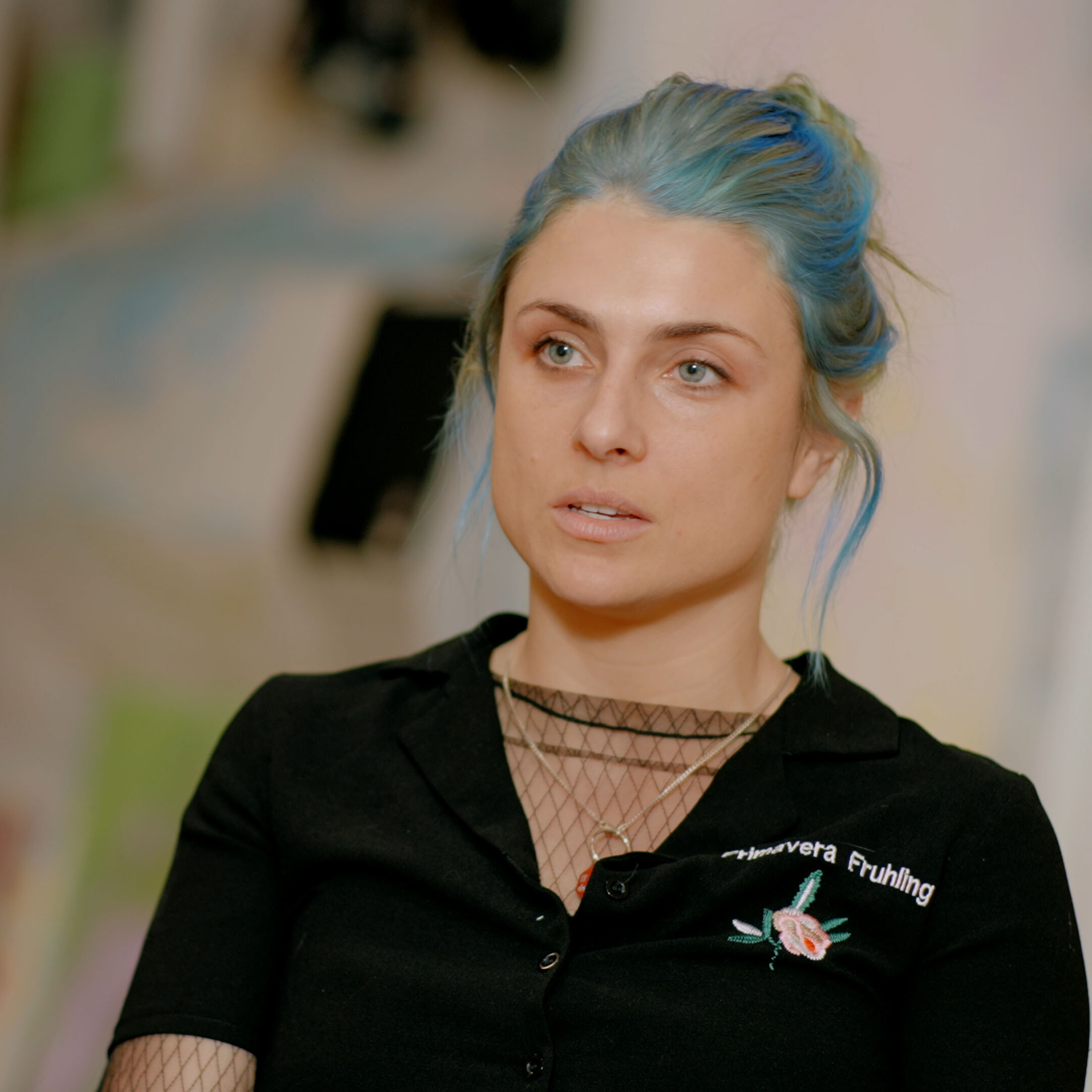 A woman with blue hair sits in front of a blurry studio. 