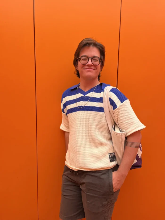 A photo of a person leaning against an orange wall with a white shirt and bag. 