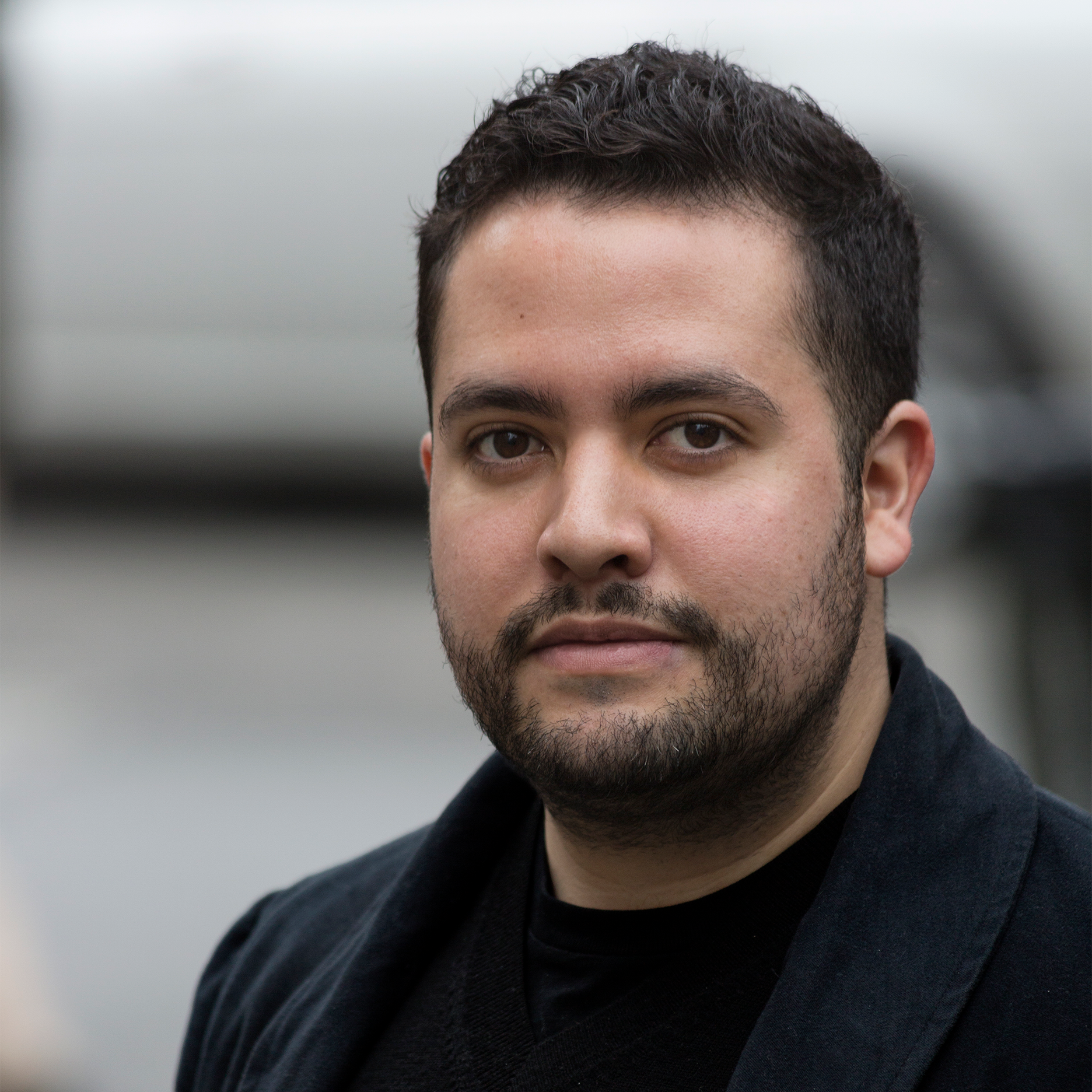 A man with a short beard and dressed in all black stands with a blurred background.