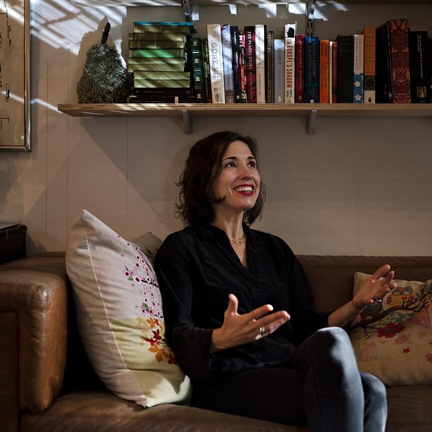 A woman sits on a couch with a book shelf behind her. 