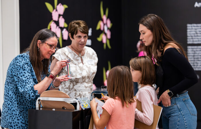Two women show a woman and two kids sensory items at a cart in an art gallery.