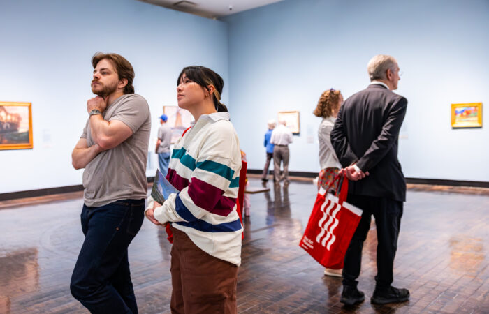 A couple look at art in a gallery, while another coupe behind them looks at different artworks with a red tote bag.