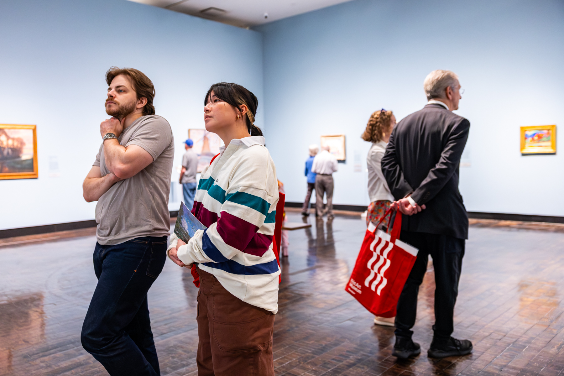 A couple look at art in a gallery, while another coupe behind them looks at different artworks with a red tote bag.
