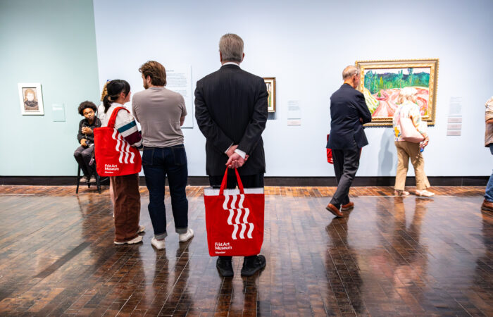 A man in a suit stands in an art gallery with a red tote bag with several other people around.