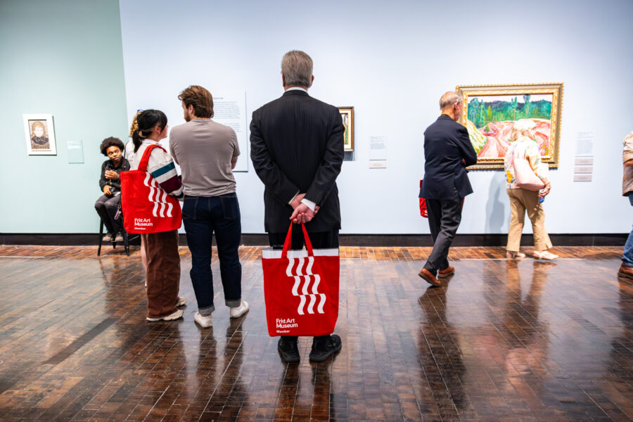 A man in a suit stands in an art gallery with a red tote bag with several other people around.