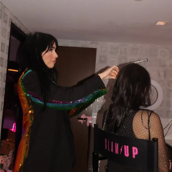 A woman in a sparkly coat does another woman's hair in a chair that says Glam Up.
