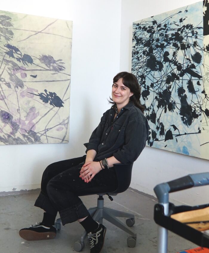 A woman sitting in an art studio with paintings of plants in the background. 
