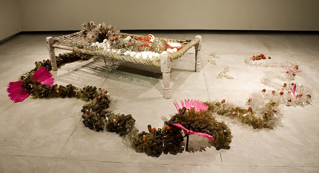 This is an installation on the gallery floor. There is a raised platform and around it, arrangements of objects. The materials in the work: Light bulbs, pigeon feathers, steel carbon wire, copper, silk thread, glass beads, cowrie shells, trim, wedding sari, seed beads, wood, horn, fleece. It measures 38 x 34 x 23 in.
