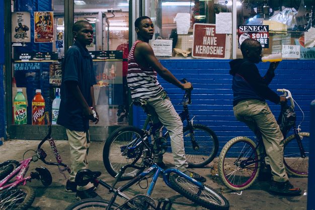 Three Black boys on bikes in front of a convenience store with a blue brick façade. The boy on the left is looking off into the distance, the boy in the middle is looking directly at the camera, and the boy on the far right is drinking juice while looking straight ahead.