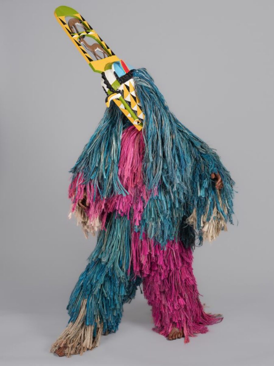 figure dressed in long strand of blue and pink jute like material with a tall painted wood mask with a beak like nose with two deer stacked on top