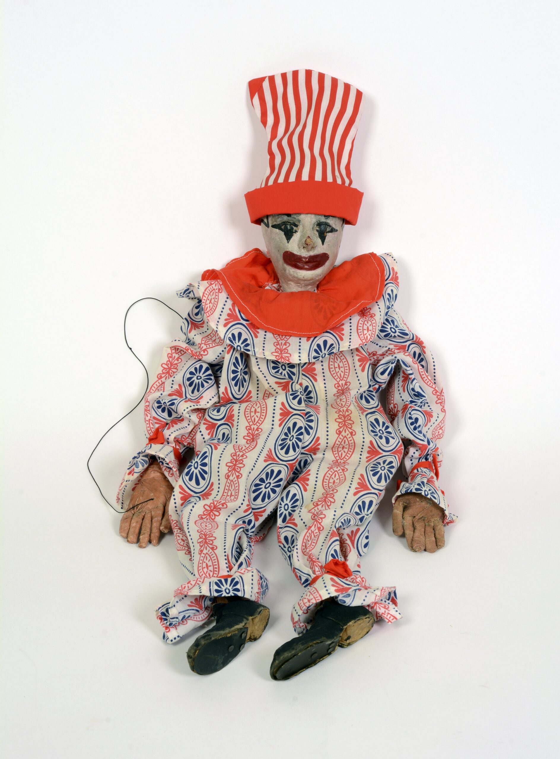A clown puppet with a painted face wears a baggy red, white and blue suit with a large, ruffled collar. A red and white striped top hat completes the ensemble.