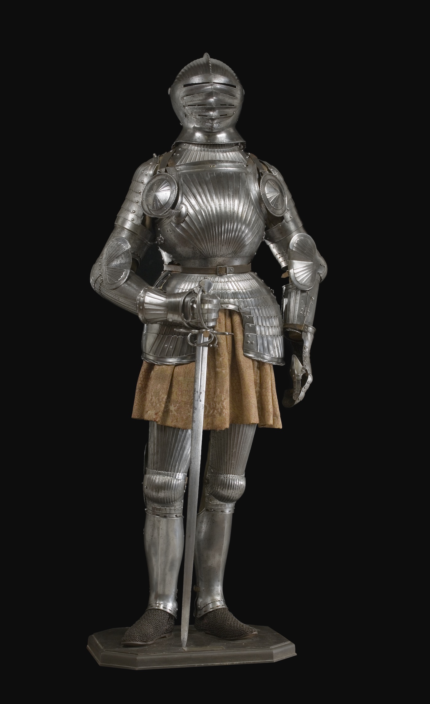 Knight statue in silver armor with a gold chainmail skirt. One arm props up a sword by the handle with the point resting on the ground by his feet.