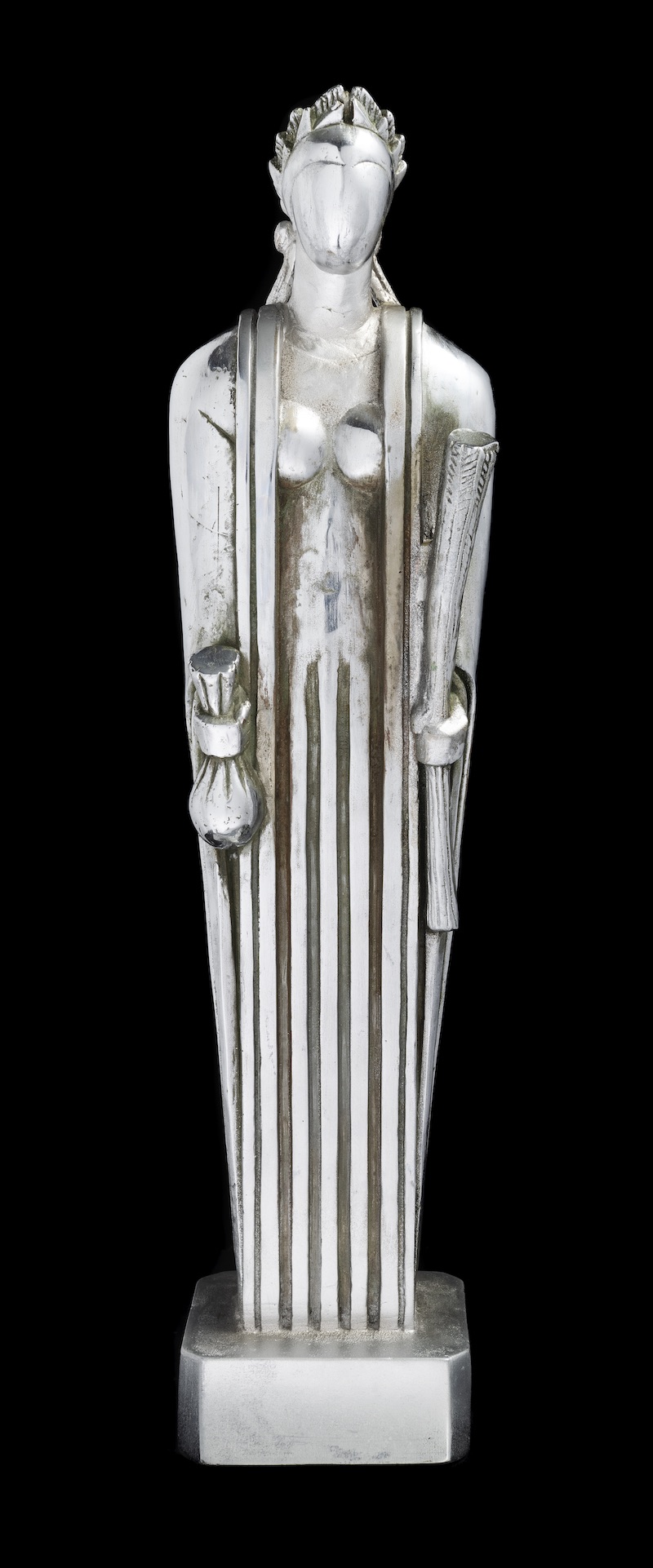 Faceless, silver colored statue of a robed, seemingly bare breasted woman holds a bag in one hand and a rolled object in the other. The upper torso melds vertical lines as the figure descends into parallel vertical lines.