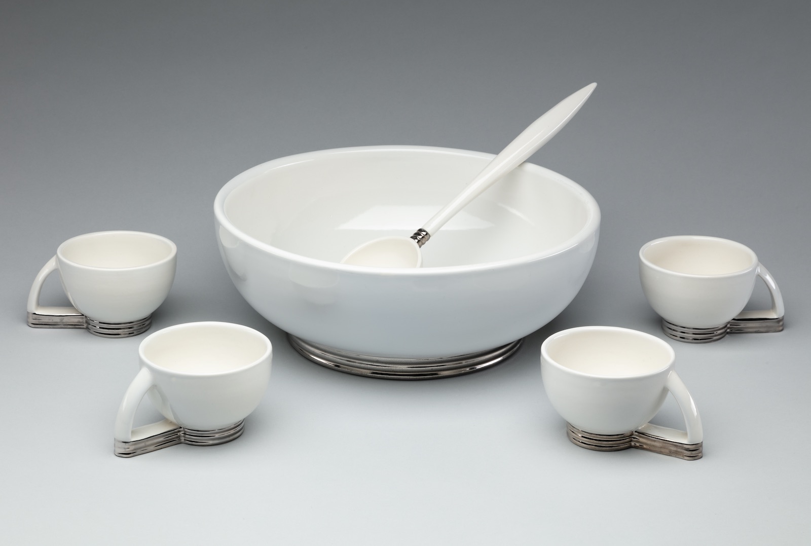 White ceramic bowl sits atop a silver bottom. The long-handled ladle rests on the bowl’s rim.  Small white cups with matching silver bottoms surround the bowl.