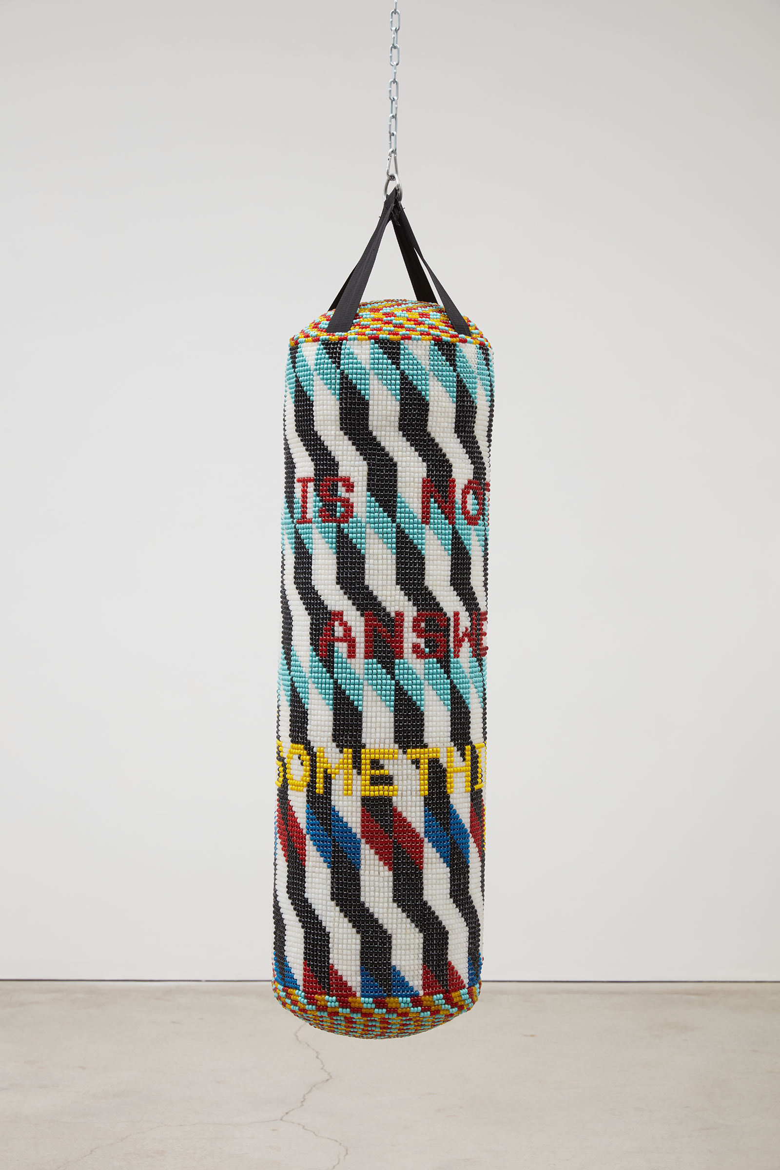 A multi-colored and beaded punching bag hangs from the ceiling against a white wall