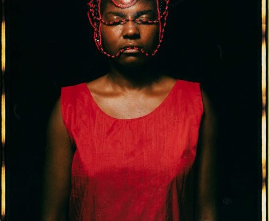 African American woman standing with her eyes close dwith red and black strings of bead draped over her head, wearing a red shirt.