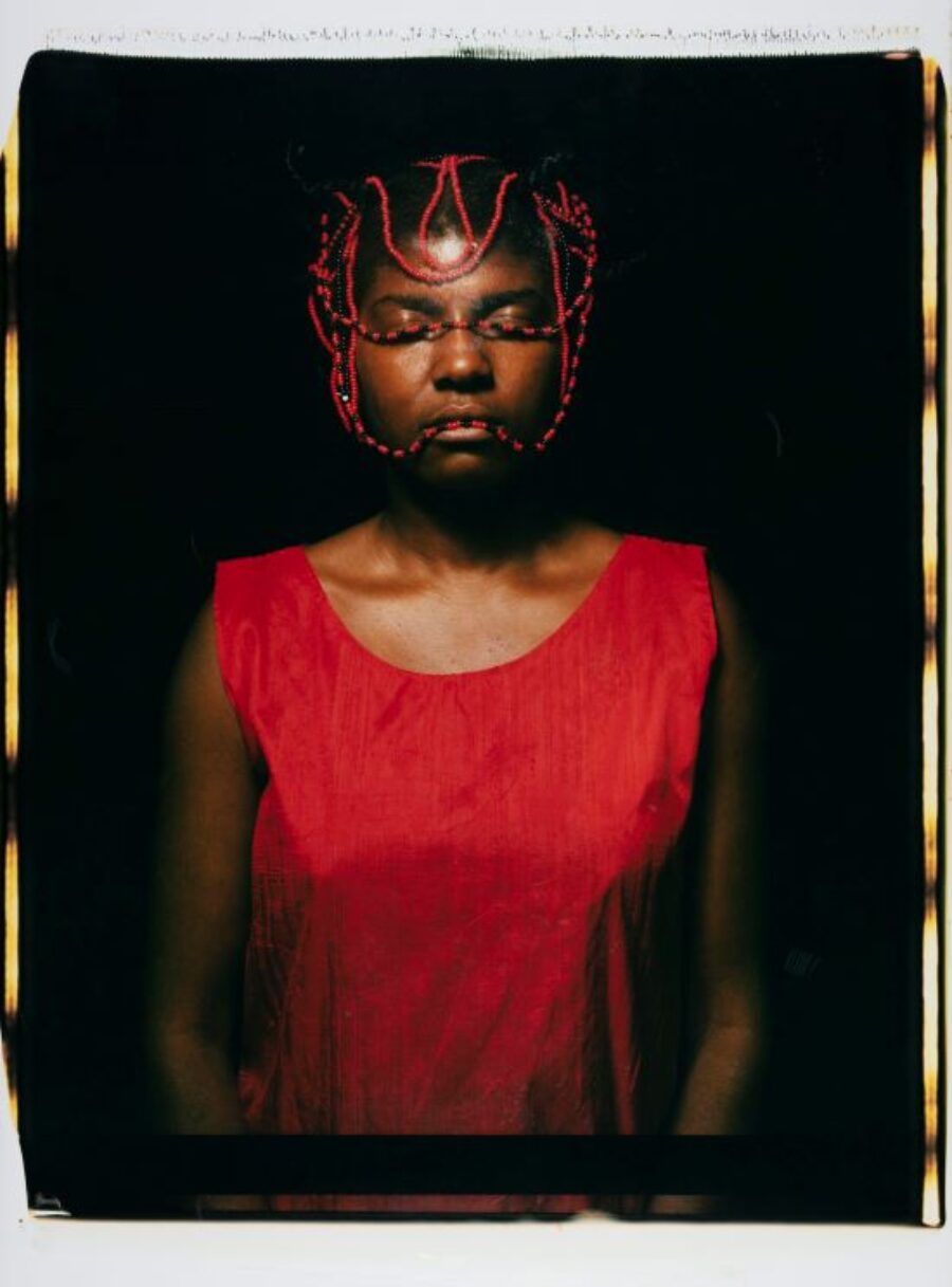 African American woman standing with her eyes close with red and black strings of bead around her head and wearing a red shirt.