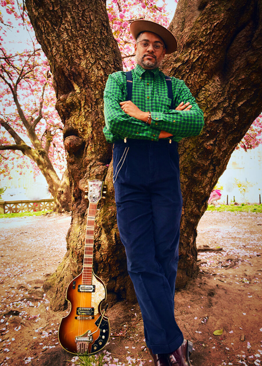 Don Flemons standing next to a tree with his guitar next to him