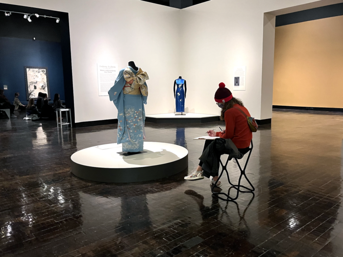 Young woman sits in Weaving Splendor gallery drawing in front of a light blue kimono