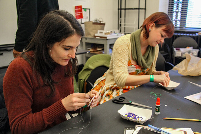 Two women sitting at a table working on an art project using thread and yarn