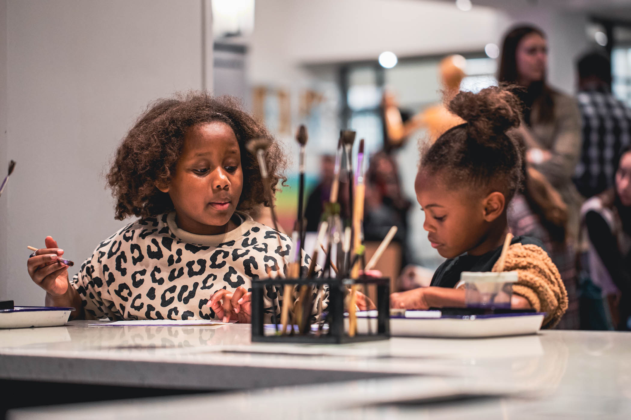 Two young girls sitting at a table with a rack of paint brushes in the center working on an art project