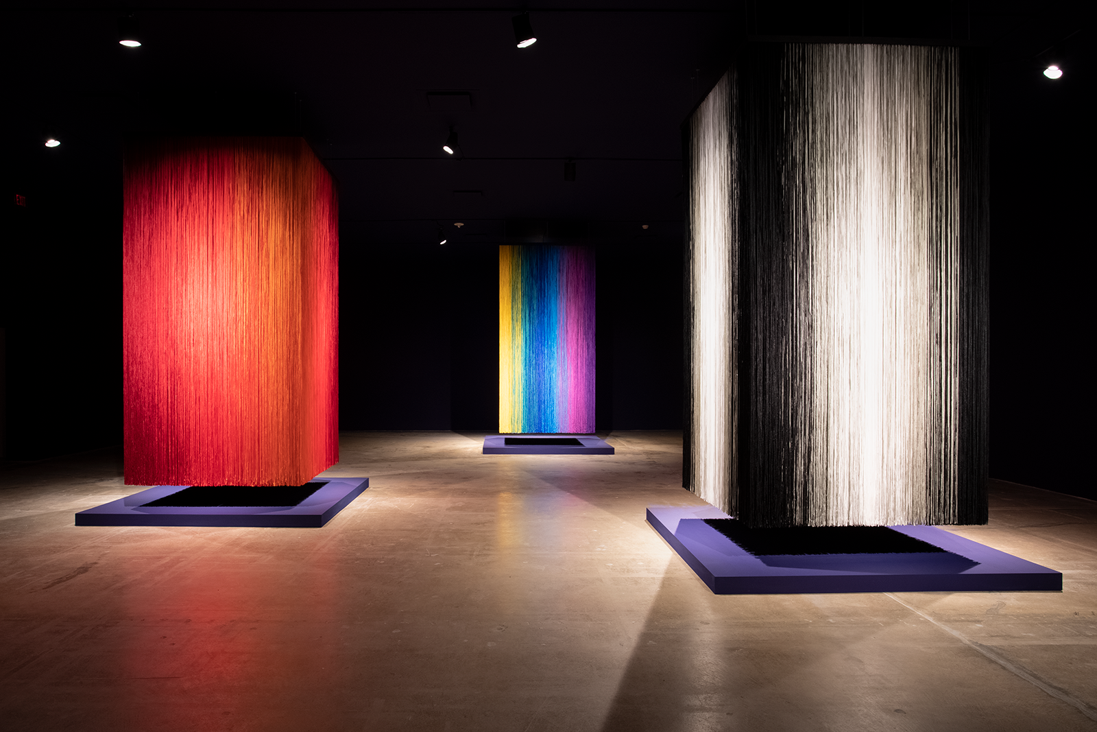Three cube-shaped sculptures made of different colors of shawl fringe handing from the ceiling