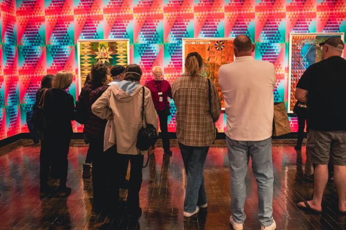 The backside of a group of people as they listen to a tour guide in a colorful gallery