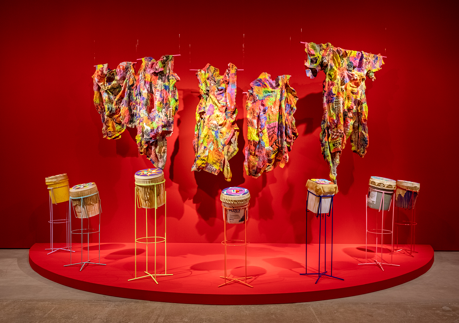 Bright red wall with seven drum-like structures and five multicolored pieces of clothing hanging above them