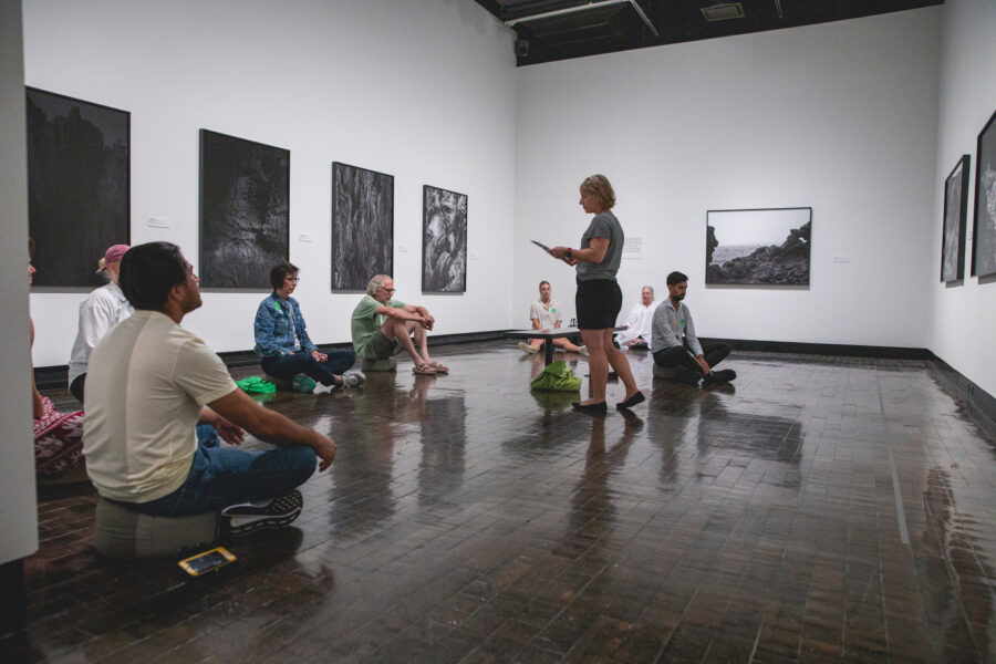 Group of people sitting on the gallery floor, meditating, led by a standing woman