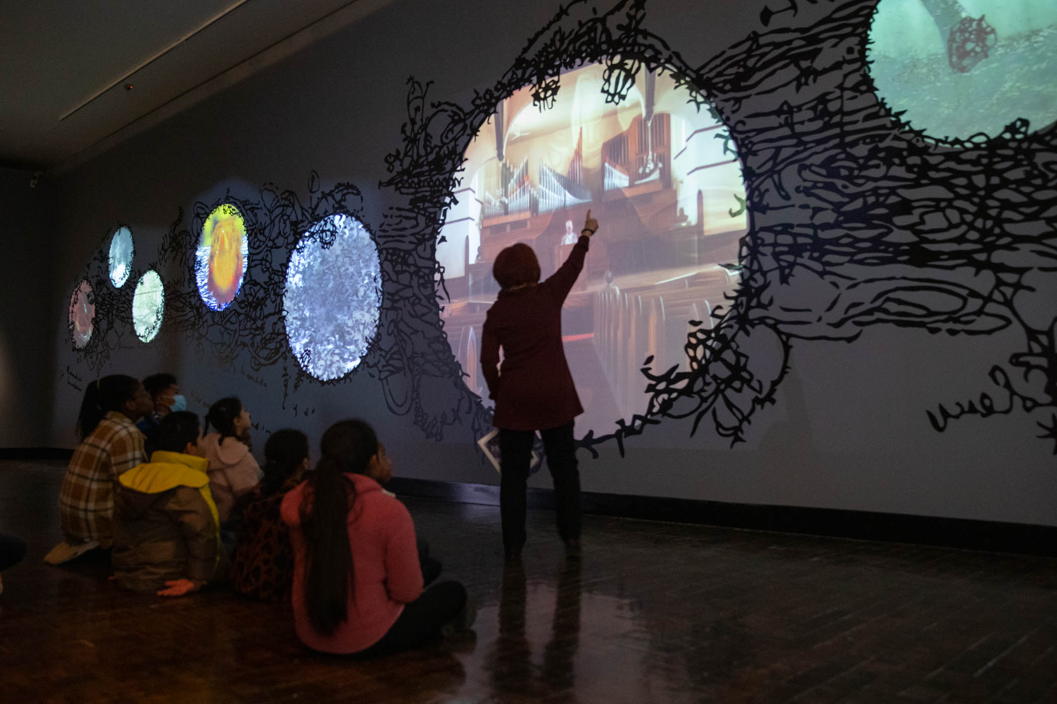 Docent pointing to a video wall with group of students sitting on the floor