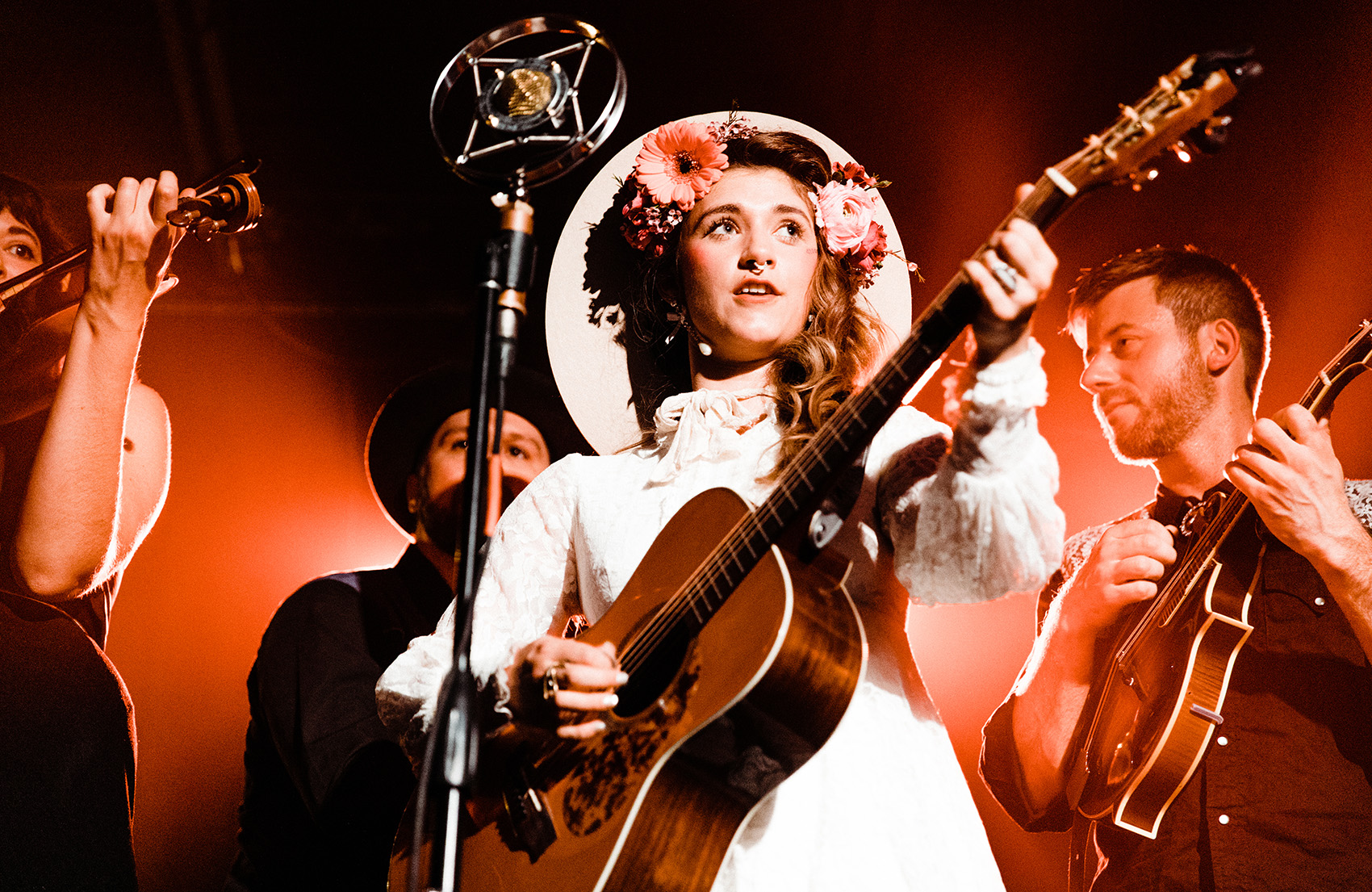 Woman dressed in a white dress with a hat and pink toned flowers in her hair playing guitar with band members behind her.